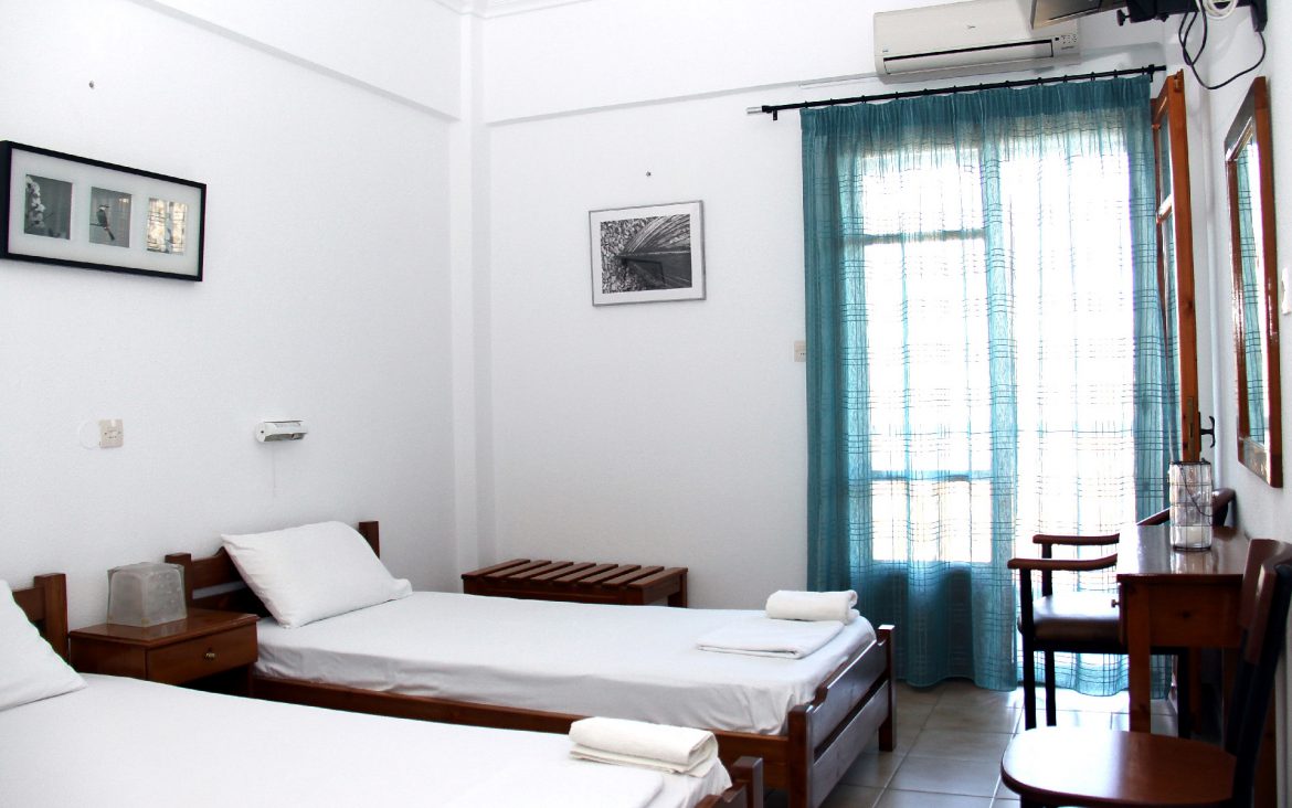 A twin room with white walls, two single beds and a balcony door behind teal curtains